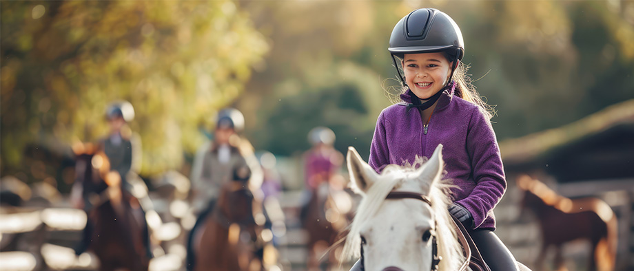 Fid'Equin : Photo d'une enfant de sexe féminin sur un cheval blanc dans une carrière d’entraînement. Elle est souriante et porte l'équipement recommandé pour un cavalier. Elle se trouve à droite de l'image. L'arrière fond est légèrement flou.